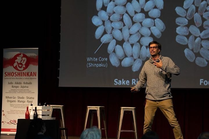 Presentador en escenario durante una masterclass sobre sake, con fondo de proyección mostrando granos de arroz.