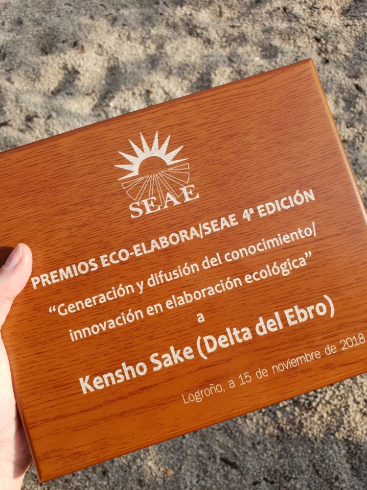 premios eco-elabora Placa del premio Eco-Elabora 4ª edición otorgado a Kenshō Sake, sostenida por una mano sobre arena.